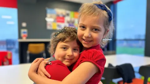 BBC Lacie (left) and Betsy (right) at school. Both in red polo shirts and have short hair with headbands. 