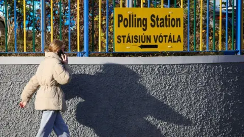 Getty Images A woman in a beige coat and grey trousers walks past a yellow sign on a fence that says 'Polling Station' in English and Irish.