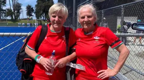 Rae Ellis Twin sisters both aged 66, with silver hair, wearing their welsh jersey shirts outside on a playing field. 