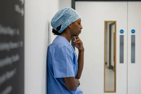 Getty Images An NHS hospital worker wearing scrubs and a hair net stands against a wall inside a hospital in Newcastle while looking pensive. Stock photo illustration.