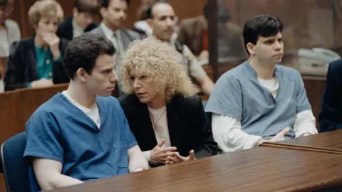 Getty Images The Menendez brothers, wearing different shades of a blue prison uniform, sit at a table with one of their attorneys during their trial in 1994. Inside the courtroom, their attorney is a woman with curly hair and appears deep in conversation with Erik Menendez. 