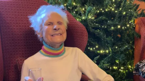 BBC Molly, who is wearing a rainbow coloured jumper, is sitting in a red armchair and smiling. She is holding a champagne glass.