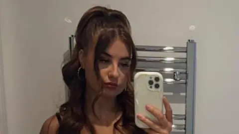 Paige Roberts Paige Roberts taking a selfie in a mirror, she has long wavy dark brown hair and large hooped earrings. Her iPhone is visible and behind her is a vertical, wall-mounted bathroom radiator 