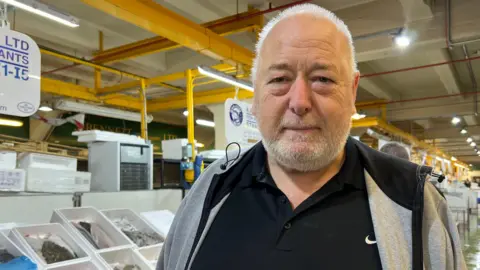 BBC Tony is looking into the camera, wearing a black top and grey hoodie. He has short white hair and a white beard. He is stood in front of trays of raw fish in an aisle at Billingsgate fish market. 