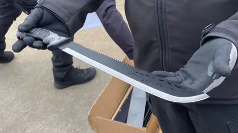 Ben Schofield/BBC A large knife with a blade that is sharpened on one side and serrated on the other. It is being delicately held by two hands wearing heavy-duty black safety gloves. In the background is a cardboard box containing smaller black boxes; it is sitting on a concrete surface. Another person wearing black boots and trousers appears to be reaching into the box. 