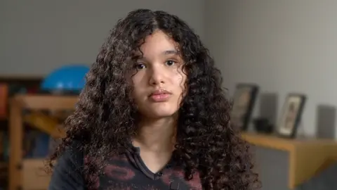 BBC Mia looks into the camera with a neutral expression. She has curly dark coloured hair that just passes her shoulders. She is wearing a dark coloured top. In the background is living room furniture and photographs are propped on one side. 