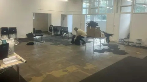 An empty shopping unit with half of the tiled flooring lifted up and water marks visible underneath. In the background there's a person lifting a tile from the floor.