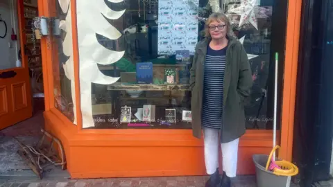 Jayne Coleman wearing a striped t shirt and green coat, stood outside her orange-painted shop front with a mop and bucket beside her