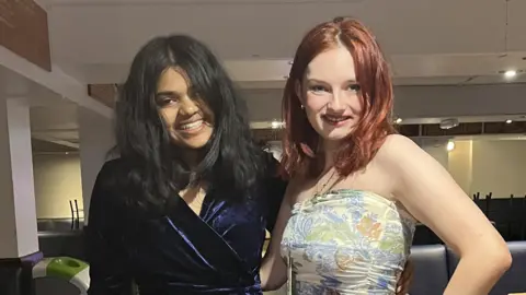 DWAS Vanya and Hannah standing together smiling at the camera. The girl on the left has dark hair and is wearing a dark blue, velvet wrap dress. The girl on the right has red-coloured hair and is wearing a cream dress with blue, green and orange floral pattern.