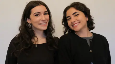 DWAS Two girls smiling at the camera against a white background. The girl on the left has long, dark, curly hair and is wearing a black long-sleeved top and silver jewellery. The girl on the right has shoulder-length, dark curly hair and is wearing a black cardigan and grey top. 