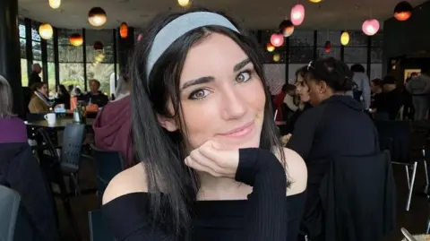 DWAS Juliette is smiling at the camera, resting her chin on her hand. She has long, dark straight hair and is wearing a blue headband and black long-sleeved, off-the-shoulder top. In the background is a restaurant with people sitting at tables and coloured lights coming down from the ceiling. 