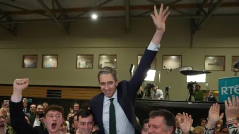 PA Media Simon Harris celebrates at a count centre. He is wearing a blue jacket and blue tie and white shirt. 