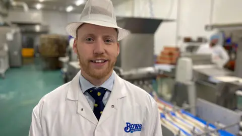 A man stands in a factory smiling at the camera. He's wearing a white hat and a white coat.