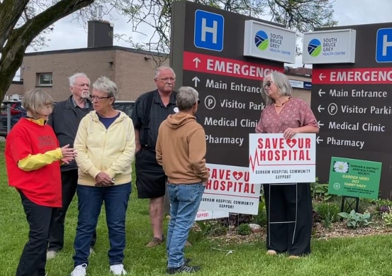 People stanidng in front of a hospital, one has a sign that says "Save Our Hospital"