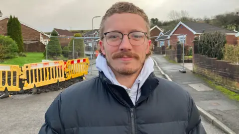 Callum Morgan stands outside the cordon. He is wearing a black winter coat with a white hooded top. He has glasses, blonde hair and a moustache and beard.