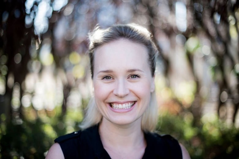 A blonde woman smiles for the camera. There is a blurry, green background behind her.