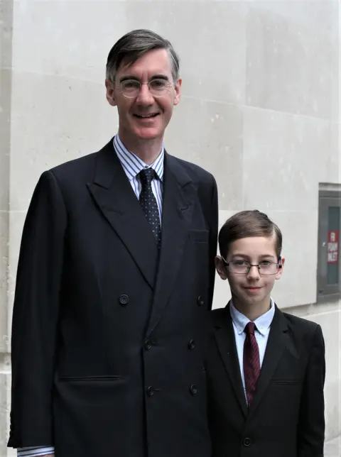 Getty Images Jacob Rees-Mogg with his eldest son, Peter Theodore Alphege in 2018. They are both wearing black suits, shirts and ties. Both also wear glasses.