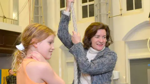 Arts Council of Wales Eluned Morgan holding a rope during a circus training session at NoFit State Circus. Eluned Morgan is stood wearing a grey cardigan and a white jumper, in front of a young girl in an orange top.