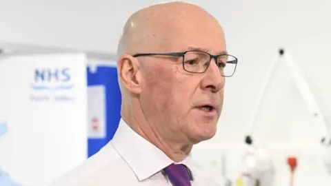 PA Media First Minister John Swinney standing in front of an NHS forth valley sign. He is bald and wearing glasses. He has a white shirt on and a purple tie.