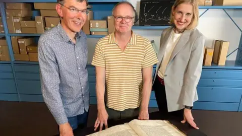 National Archives Tim Thornton, wearing a blue shirt, jeans and glasses, stands with actor Jason Watkins, in a yellow striped top and glasses, and presenter and historian Tracey Borman, wearing a grey blazer and with blonde hair.