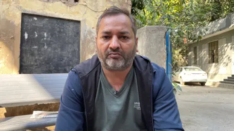 A man in a grey V-neck shirt and an unzipped blue coat stares directly at the camera while sitting in a residential area. A white car is seen over his left shoulder.