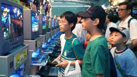 Getty Images Several children gather around a series of screens, with one holding a controller and playing a video game. The PlayStation console is attached to the screen and outputting a game. It is an old CRT screen.