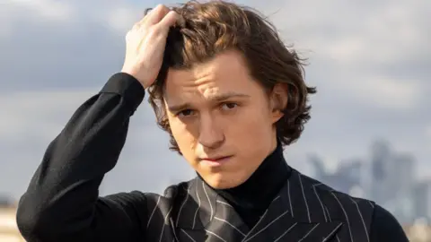 Getty Images Tom Holland wearing a sharp pinstripe black suit runs his fingers through his hair and stares intently at the camera.