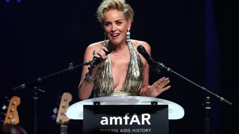 Getty Images Sharon Stone, wearing a silver dress and speaking into a microphone, at Cinema Against AIDS Gala in France in 2014