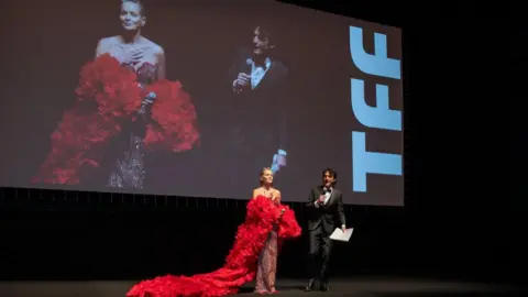 Getty Images Sharon Stone in a dress with a long red train, speaking in front of a screen showing herself, at the Torino Film Festival, November 2024