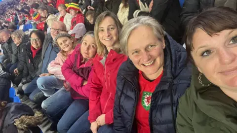 Ruth Billingham A selfie of fans at the football. Ruth is on the far right and is taking the photo, she is wearing a green jumper and has gold hoop earrings and is smiling. A woman is sat next to her and is wearing a red Wales shirt, she has grey hair and is also smiling. Next to her is a woman with blonde hair and a pink jacket. She is next to another woman with a pink jacket. Next to her is a girl with a pink Adidas coat, who has her arms crossed and is smiling at the camera. Next to her is an older couple who are both smiling at the camera.