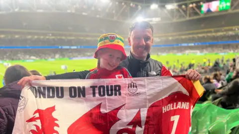 Joey Healey Joey Healey and Harlow supporting Wales in Dublin. They are holding a Wales flag, with the stadium behind them. Harlow has a Wales bucket hat on and a Wales football top on.