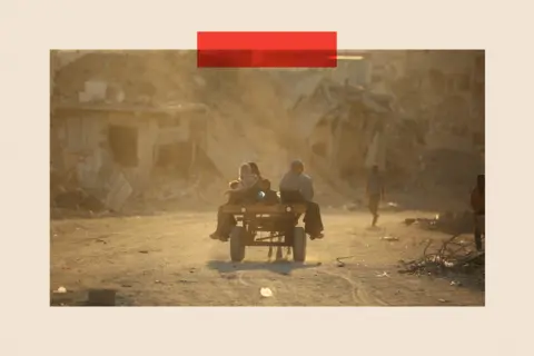 Getty Images A group of people sit on a wooden cart pulled by an animal, surrounded by rubble and ruined buildings.