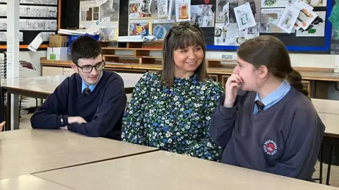 Munaza Rafiq / BBC Two pupils sit with a teacher at a High School. They are in a classroom and work is displayed on the wall behind them.