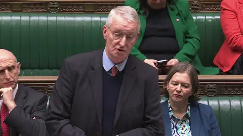 Hilary Benn speaking to the House of Commons. He is wearing a grey suit, blue shirt and red tie. He has glasses and grey hair. Behind him are other members of parliament on green leather seats.