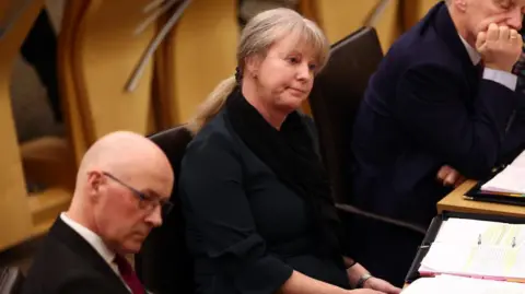 Getty Images Shona Robison, wearing her hair in a ponytail and a black scarf and jackets sitting in the Scottish Parliament chamber. To her left is John Swinney.