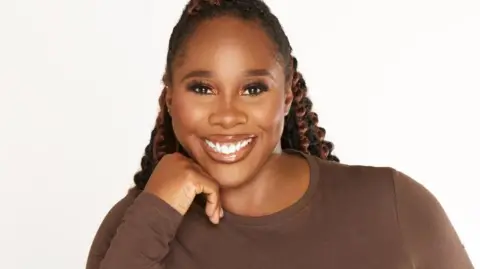 Amanda Akokhia Denise  is wearing a brown top and is pictured against a plain white background. She has long, black braids with curls and has her chin resting on her hand and is smiling.