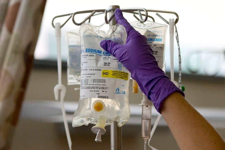 A gloved hand hooks a bag of clear fluid to a pole.