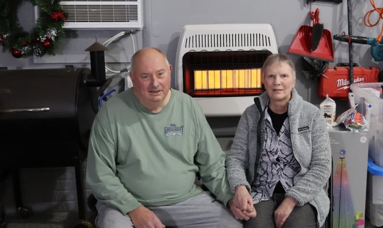 A man and woman sit side by side in a garage next to a heater, smiling and holding hands.