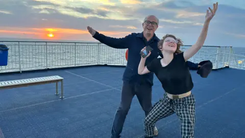 Julian Frances Julian Frances and April, smiling and throwing their arms in their air on board a ferry at sunset. Mr Frances wears black-rimmed glasses, a navy shirt and jeans. April wears glasses, a black t-shirt and chequered trousers, holding a phone in one hand.