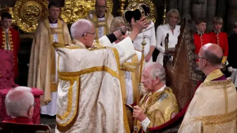 Reuters Justin Welby, in white and gold robes, holds a crown over King Charles' head as he sits on a throne.