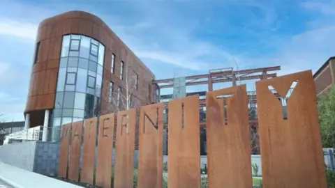 Belfast Trust The new maternity hospital at the Royal Victoria site. There is a wooden fence with maternity on it in front of a modern wood and glass building.