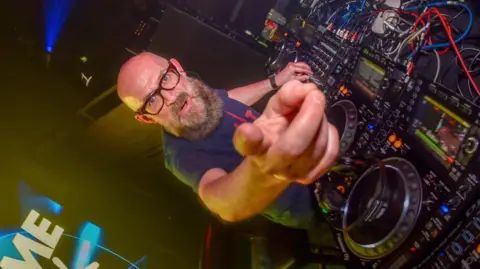 Dominic Simpson DJ Graeme Park wearing glasses and with a beard points at the camera while on the decks