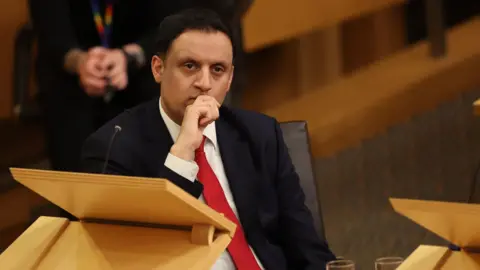 PA Media Anas Sarwar sitting behind a desk in the Scottish Parliament chamber. He has his right hand to his mouth and is wearing a black suit, white shirt and red tie.