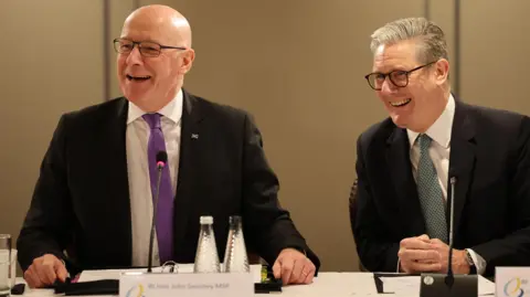 EPA John Swinney and Sir Keir Starmer laughing while sitting at a table at the British-Irish Summit meeting in Edinburgh. Both men are wearing glasses, black shirts, white shirts and ties. There are microphones and bottles of water on the table in front of them.