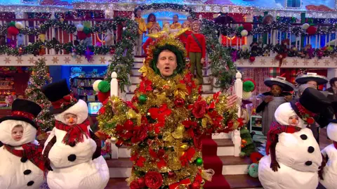 RTE Paddy Kielty covered in Christmas decorations and children dressed as snowmen and others standing on stairs behind him. The stage is also decorated for Christmas