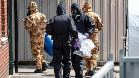 Getty Images Members of the emergency services in protective gear while searching Dawn Sturgess home in Wiltshire. Two are in black full body suits and gas marks. Two are in camouflage coloured suits and face masks.