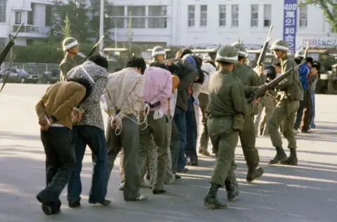 Getty Images Armed South Korean troops wearing helmets and with large guns round up protestors - mainly men, with their hands tied behind their backs, all in civilian clothes - following a raid in the city of Gwangju after an uprising against the Chun Doo-hwan dictatorship, South Korea, 27 May 1980