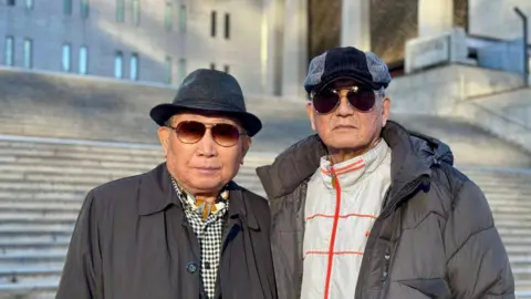 BBC / TESSA WONG Two 80-something South Korean men stand in front of the steps of a large building in Seoul