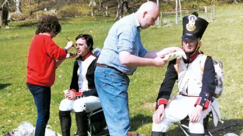 Family photo Two men, dressed in historical soldier outfits are having makeup applied to them. They are both sitting on chairs. A lady in a red jumper, with brown hair and jeans on, is applying makeup to one of the actors and a young Dave Myers is applying makeup to the other actor. He is dressed in a checked shirt and blue jeans. He has a look of concentration on his face.