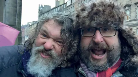 Family photo Si and Dave are taking a selfie and making funny faces to the camera. Si has his head resting on Dave's shoulder. Dave is wearing a furry hat and has glasses on. It is cold as they are both wearing winter coats and scarves.